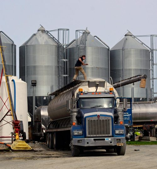 A worker loads a fuel truck in front of large industrial silos outdoors.